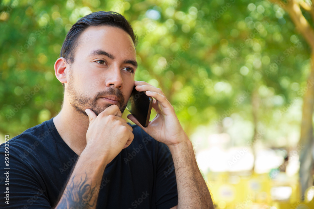 Pensive guy talking on cellphone outdoors. Handsome man with tattooed arm speaking on mobile phone on park, touching chin, looking away. Phone talk outside concept Pensive guy talking on cellphone outdoors. Handsome man with tattooed arm speaking on mobile phone on park, touching chin, looking away. Phone talk outside concept