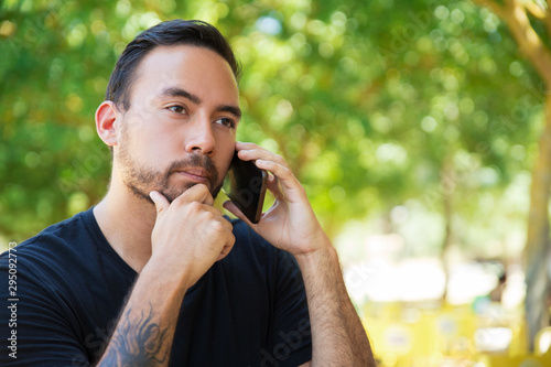Pensive guy talking on cellphone outdoors. Handsome man with tattooed arm speaking on mobile phone on park, touching chin, looking away. Phone talk outside concept