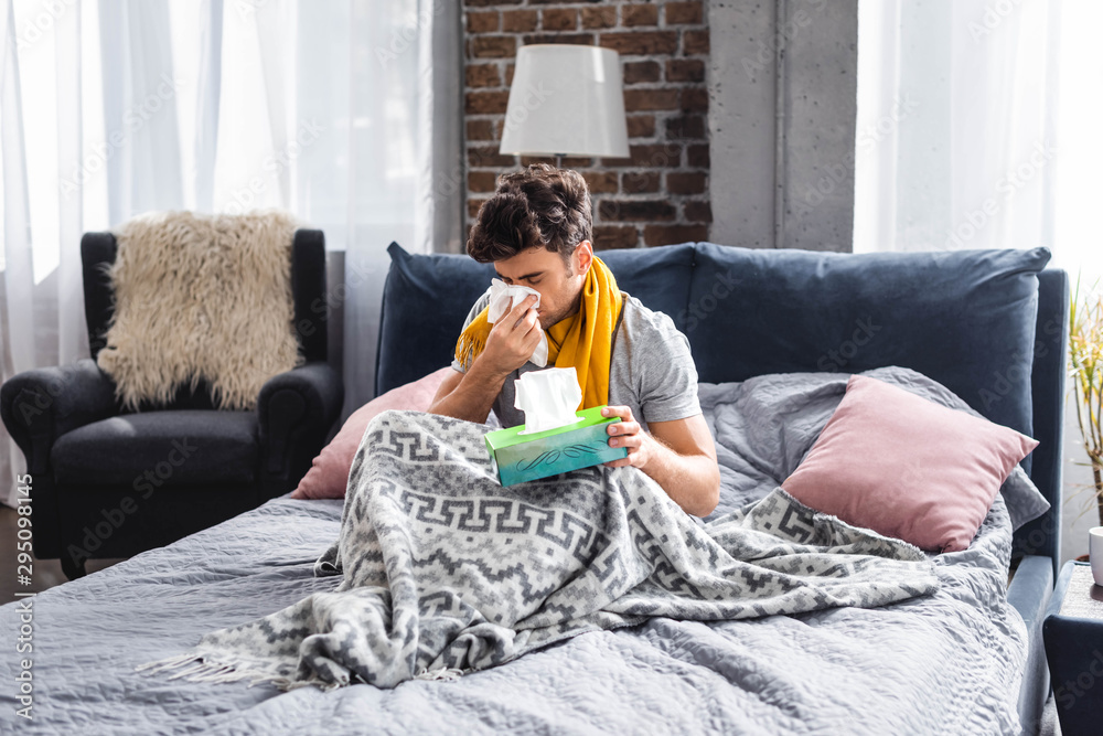 Obraz premium sick man in scarf sneezing and holding napkin in bed