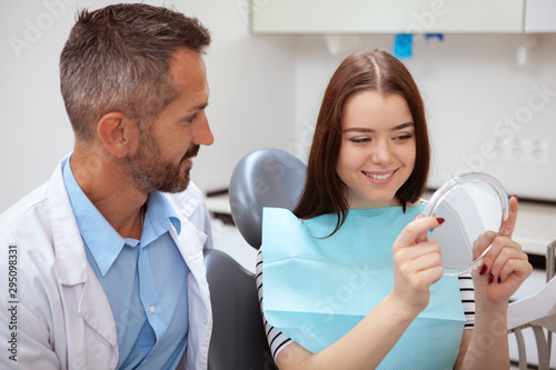 Happy beautiful woman examining her healthy white teeth in the mirror. Attractive female patient looking in the mirror after dental treatment by professional dentist