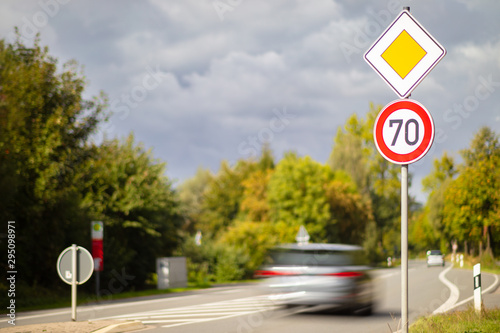 A yellow square sign on the main road and a round red and white sign with a speed limit of 70 kilometers per hour on a German country road. A car drives past high speed.