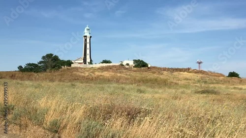 Lighthouse on a hill near the edge of the steppe, breaking off into the sea. The wind slightly sways tall grass. Static shot.