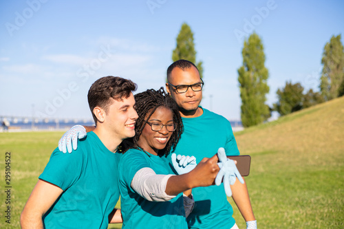 Happy united volunteer team taking selfie while cleaning lawn. Young men and woman wearing uniforms, holding smartphone, smiling at screen, posing. Voluntary society concept
