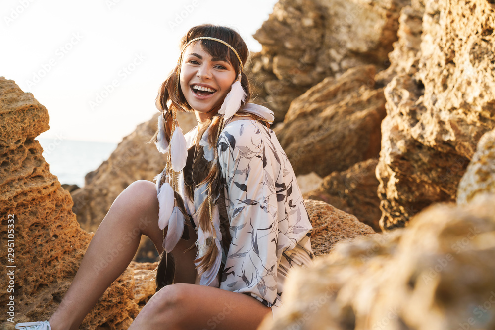 Image of happy hippy girl wearing feather headband sitting by rock