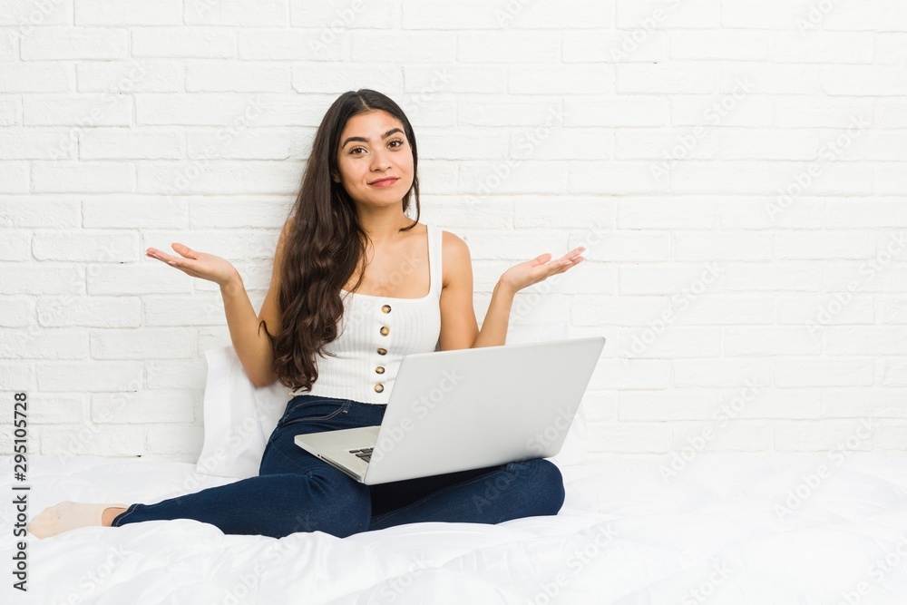 Young arab woman working with her laptop on the bed doubting and shrugging shoulders in questioning gesture. Young arab woman working with her laptop on the bed doubting and shrugging shoulders in questioning gesture.