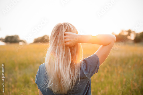 Murais de parede Young woman with blonde hair standing on the field and looking on the sunset