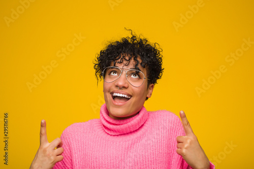Εκτύπωση καμβά Young african american woman wearing a pink sweater indicates with both fore fingers up showing a blank space