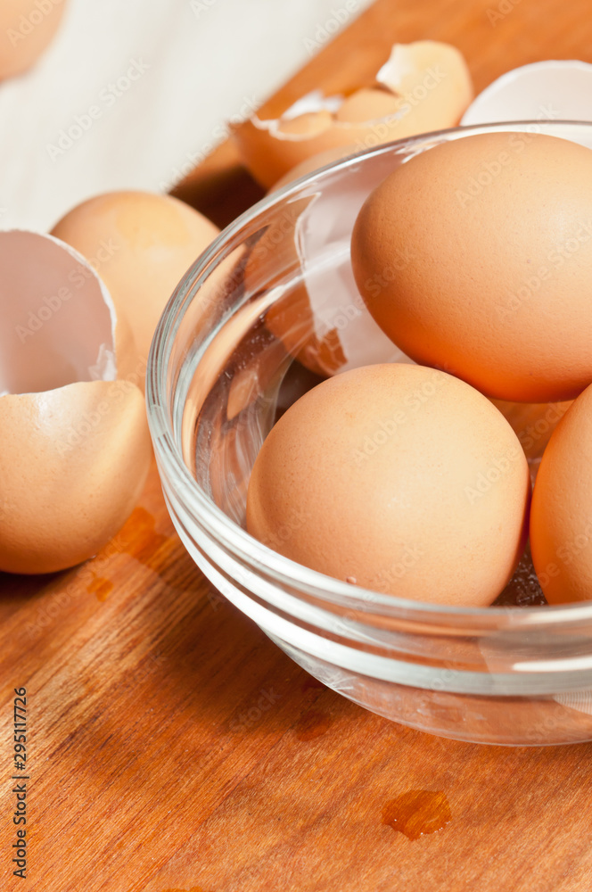 Top view, close up of a glass bowel of three, fresh, brown eggs on a wood cutting board with shells of open egg on side Top view, close up of a glass bowel of three, fresh, brown eggs on a wood cutting board with shells of open egg on side