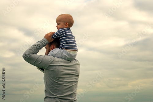 A child on his father’s neck. Walk near the water. Baby and dad against the sky.