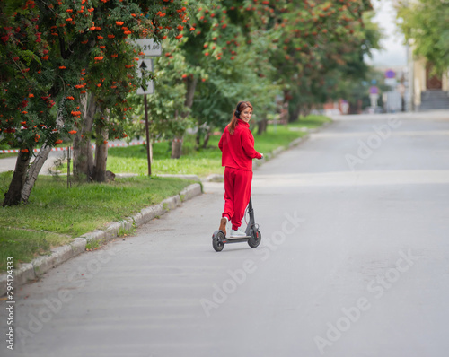 Red-haired girl in a red tracksuit drives an electric scooter. A young woman in oversized clothes rides around the city and listens to music using wireless headphones. View from the back.