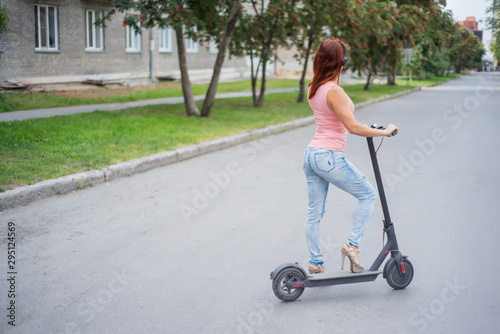 A red-haired young woman in ripped jeans and high-heeled sandals rides an electric scooter on the road and listens to music on her bluetooth wireless headphones. Convenient electric transport