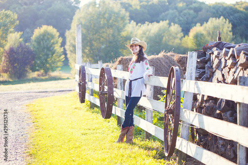 Happy young cowgirl standing at farm fence. Woman wearing blouse, cowboy hat and western boots posing at firewood and hay bales. Countryside, farming, agriculture
