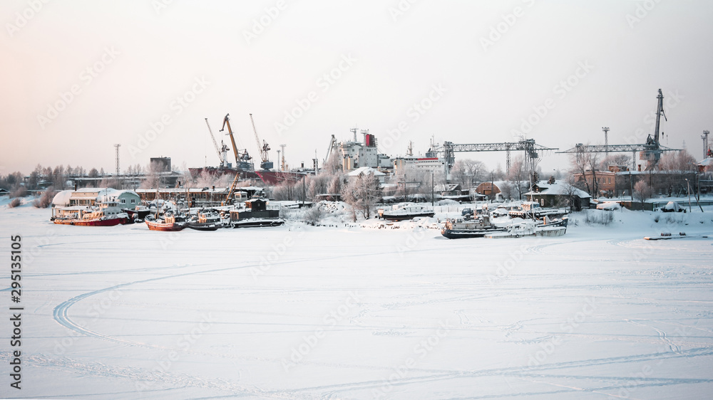 The ships, cranes and water vessels are frozen in the port. Winter cold day at the river port. Gray sky and frozen river. Everything is covered with snow