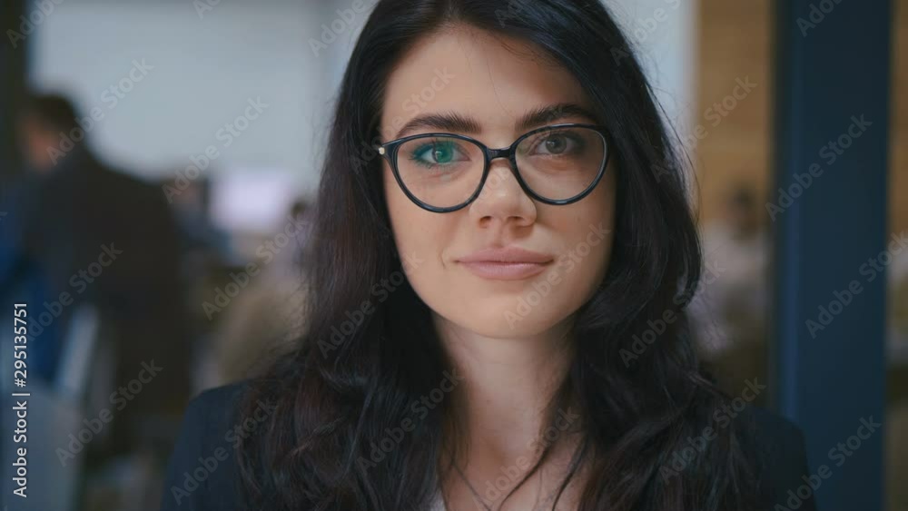 Beautiful confident young brunette woman pretty face looking at camera in office, close up portrait. Wearing eyeglasses. Office work on background. Beautiful confident young brunette woman pretty face looking at camera in office, close up portrait. Wearing eyeglasses. Office work on background.