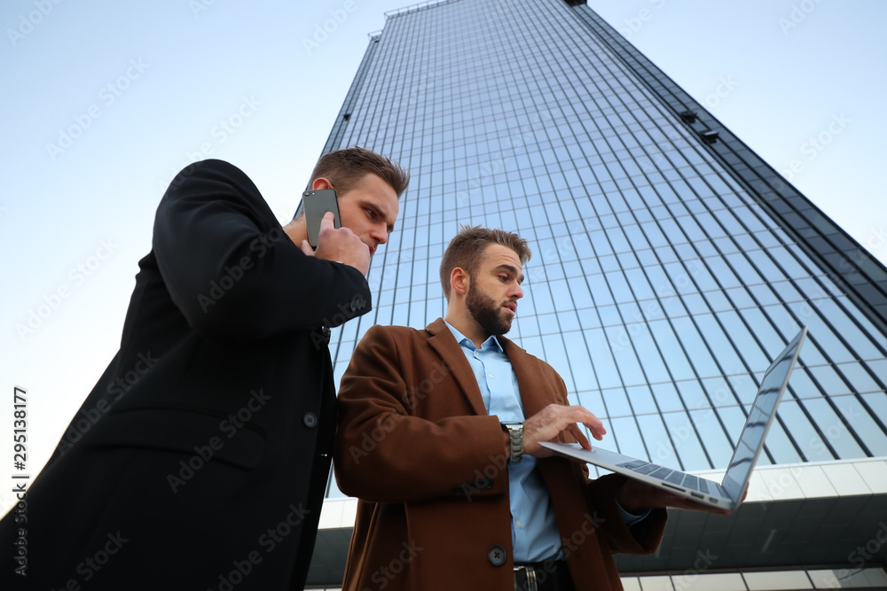 Two young guys discuss their business project and look at information on a laptop against the background of an office building
