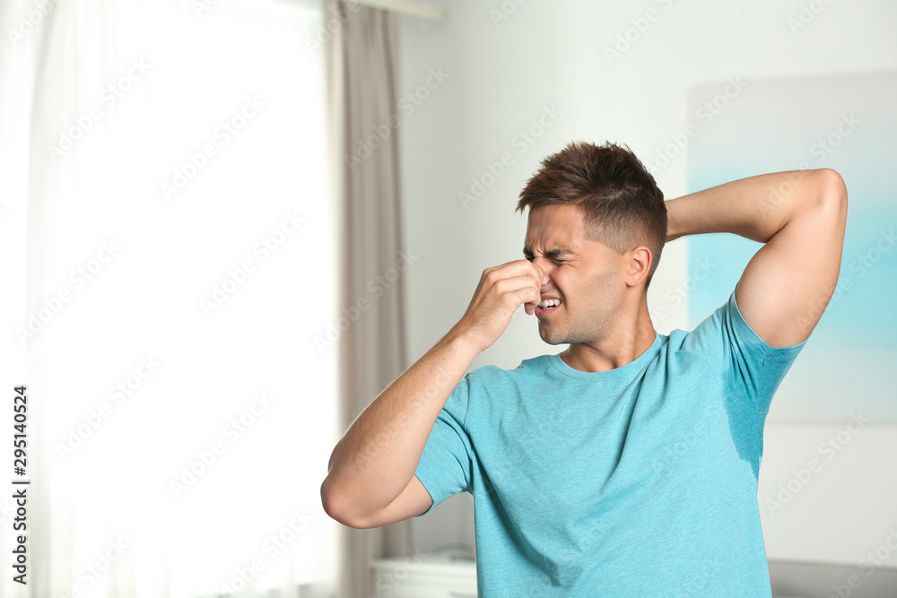 Young man with sweat stain on his clothes indoors, space for text. Using deodorant
