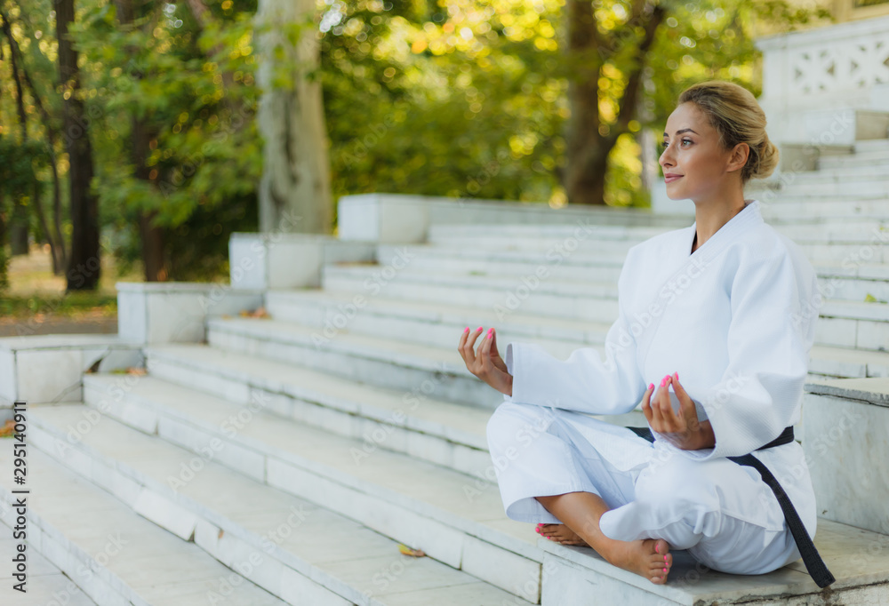 Portrait of young woman in white kimono with black belt. Sport woman sitting on stairs and meditating outdoors. Martial arts Portrait of young woman in white kimono with black belt. Sport woman sitting on stairs and meditating outdoors. Martial arts