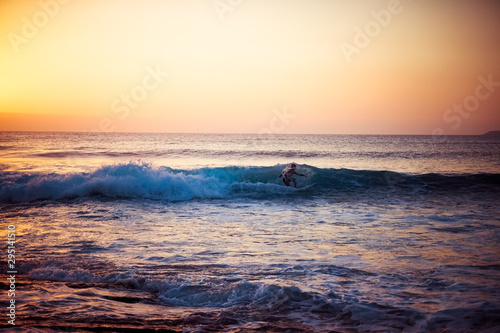 The silhouette of a surfer riding a wave at an empty surf spot. Young surfer rides the wave during sunset. Image © Manpeppe