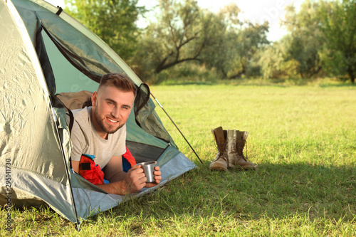 Young man in sleeping bag with cup of drink lying inside camping tent
