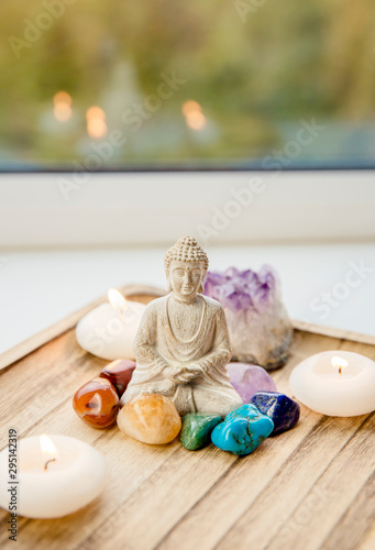All seven chakra colors crystals stones around sitting Buddha figurine on natural wooden tray. Balance and calm energy flow in home concept.