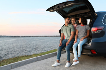 Happy young family sitting in car trunk on riverside