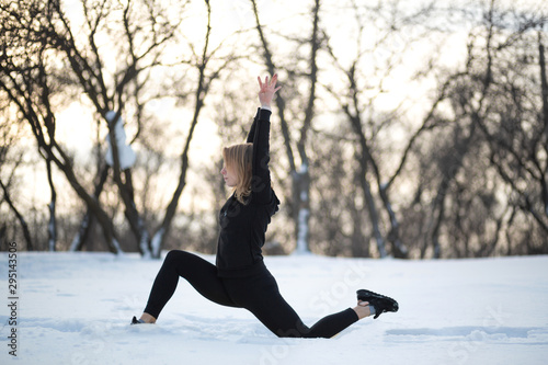 Young caucasian female blonde in leggings stretching exercise on open air in snowy forest. fit and sports lifestyle