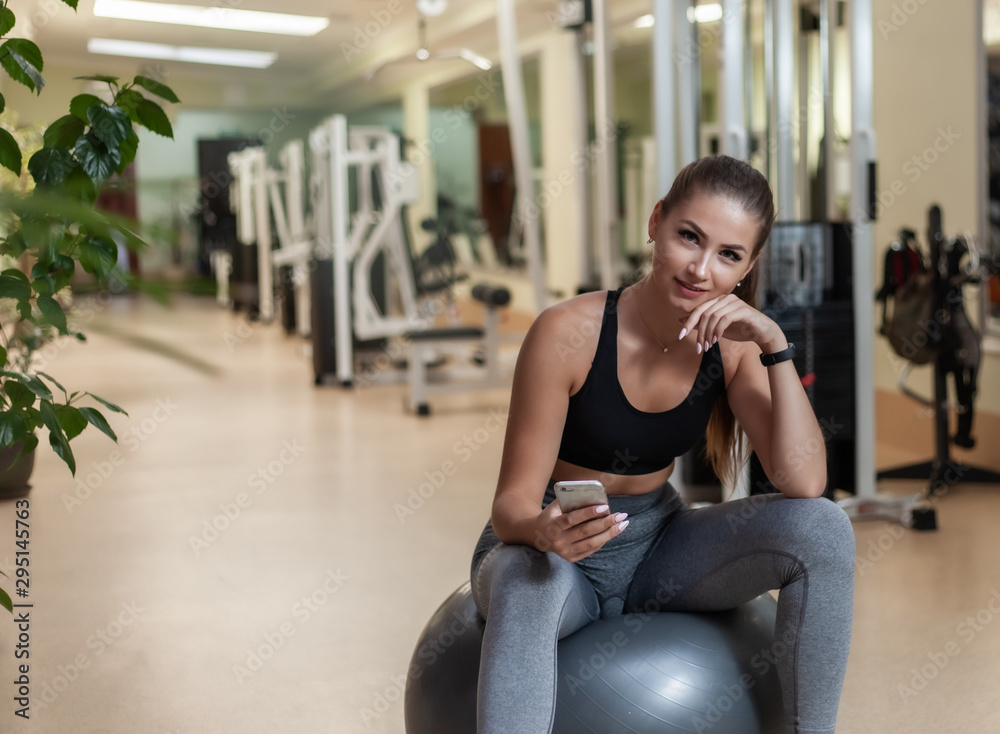 Young sport woman in sportswear using smartphone while sitting on fit ball in the gym. Workout break. Healthy lifestyle concept Young sport woman in sportswear using smartphone while sitting on fit ball in the gym. Workout break. Healthy lifestyle concept