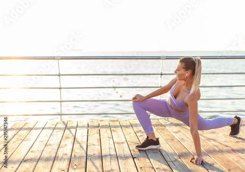 Morning workout. Warming up before training. Young fit woman in sportswear doing leg stretching on the beach at sunrise