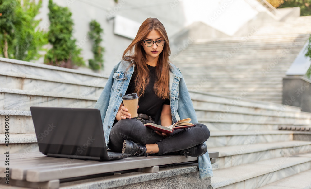 Modern students. Distance learning. Young hipster girl reads book while sitting on bench with a laptop and drink coffee Modern students. Distance learning. Young hipster girl reads book while sitting on bench with a laptop and drink coffee