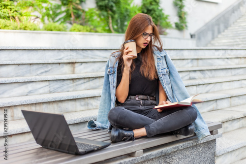 Modern students. Distance learning. Young enthusiastic woman reads book while sitting on bench with a laptop © splitov27