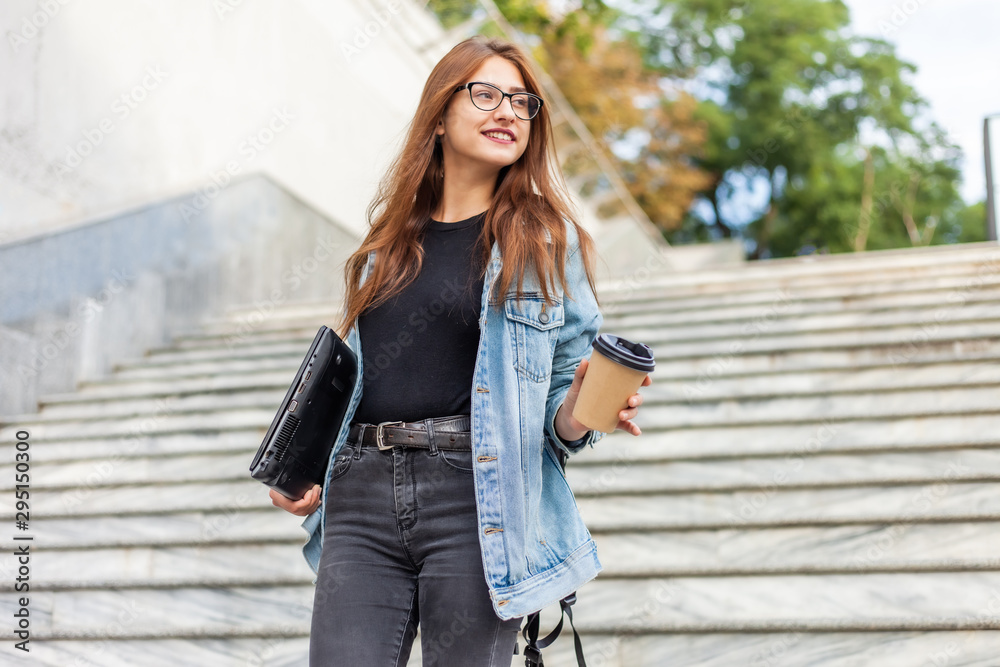 Young cheerful student woman in a denim jacket and glasses goes down the stairs with a laptop and a coffee cup in her hands in the city Young cheerful student woman in a denim jacket and glasses goes down the stairs with a laptop and a coffee cup in her hands in the city
