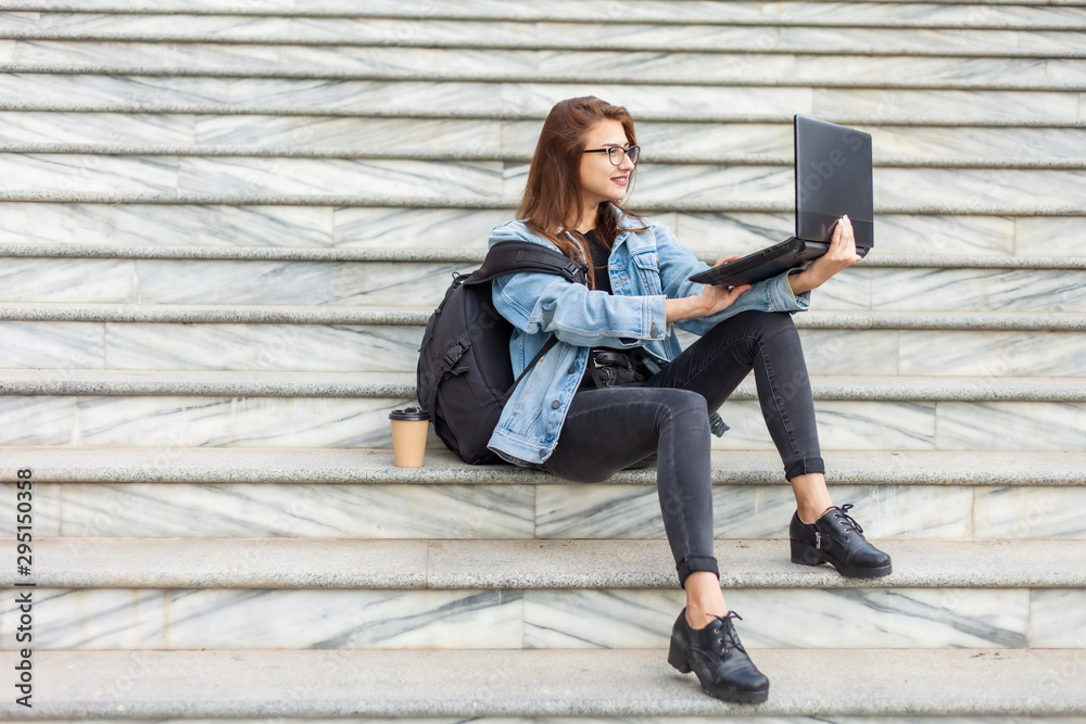 Young modern woman student in denim jacket and glasses sitting on the stairs with laptop. Watching video. Distance learning. Modern youth concept.