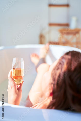 Image from back of young woman with wine glass with champagne in her hands lying in bath.