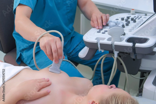 Young woman patient on ultrasound diagnostic of mammary glands on bust in clinic. Doctor runs ultrasound sensor over patient's mammary glands working on scanner panel. Diagnosis of internal organs.