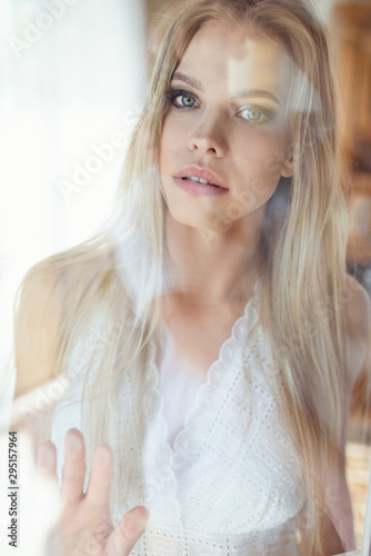 Beautiful portrait of blonde woman, with sensual look into camera. Behind the window glass.