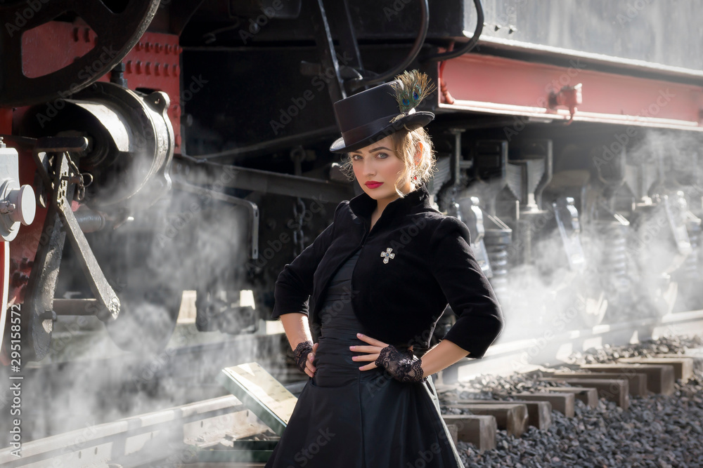 Beautiful girl in a black dress and hat near an old steam locomotive and big iron wheels. Blond beauty. Vintage portrait of the last century, retro journey.