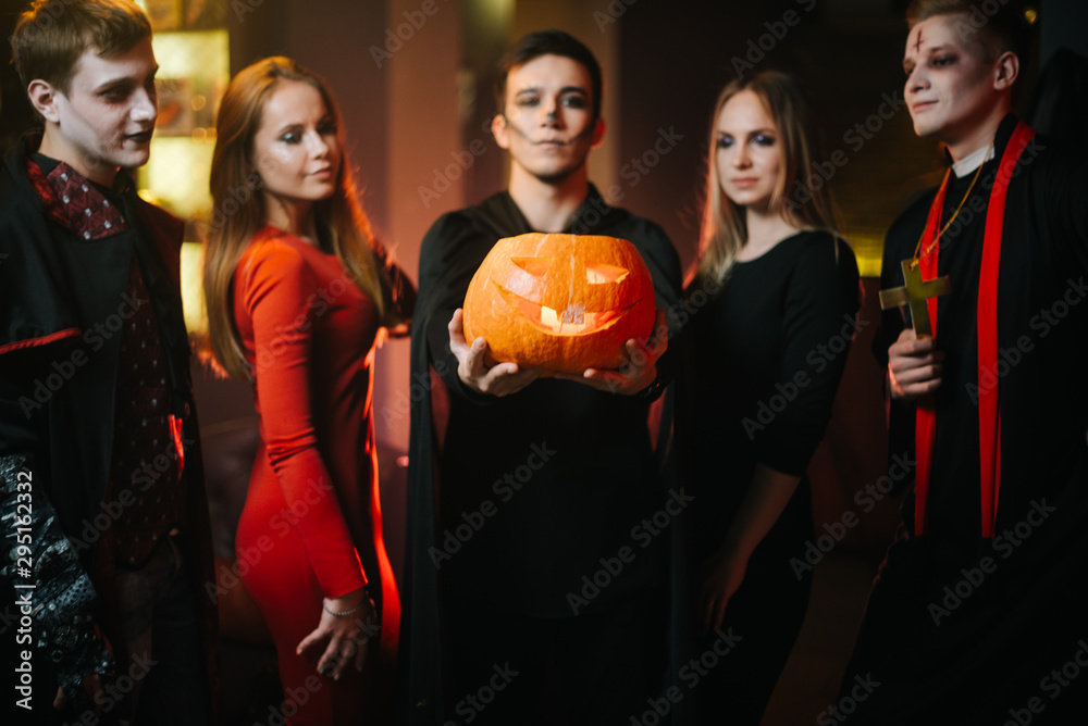 Group of friends celebrating Halloween. Guy wearing a bat costume holds a carved pumpkin on his outstretched arm. His friends look at the pumpkin Group of friends celebrating Halloween. Guy wearing a bat costume holds a carved pumpkin on his outstretched arm. His friends look at the pumpkin