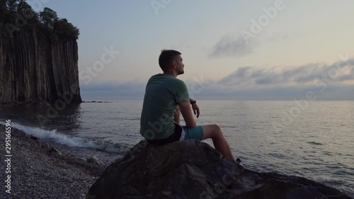 A thoughtful man sits on a rock near the sea and enjoys nature at sunset .