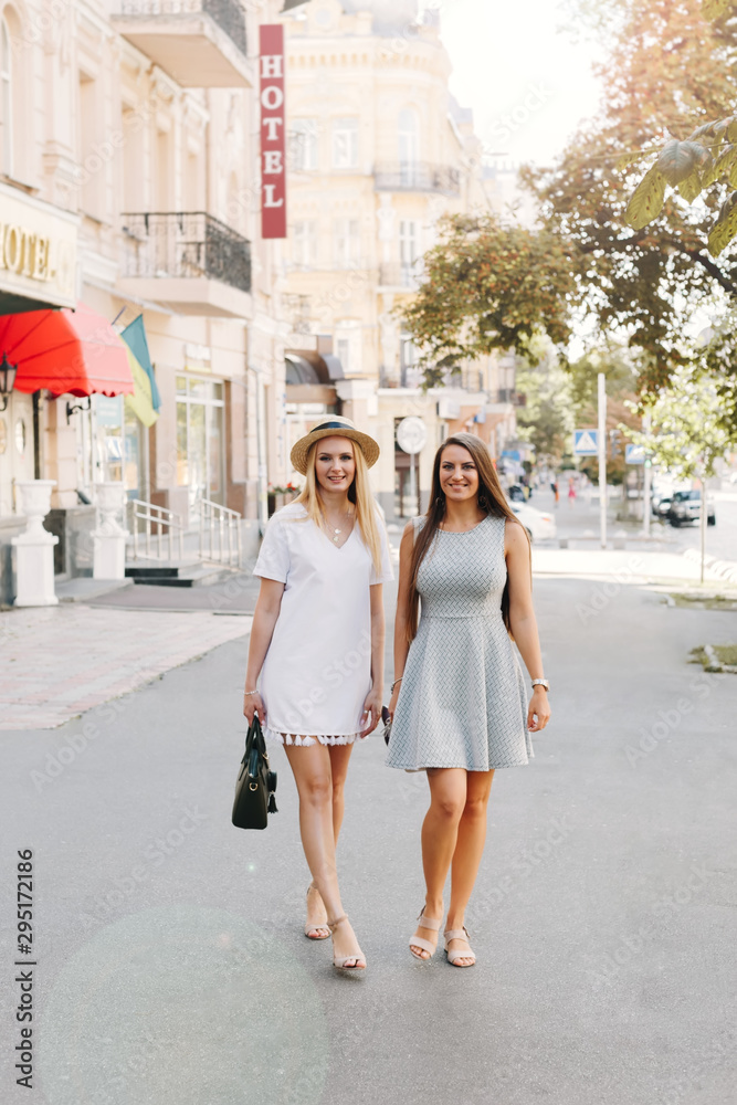 Two happy smiling girls are walking in the sunny city. Beautiful blonde and brunette walking down the street, students, travelers