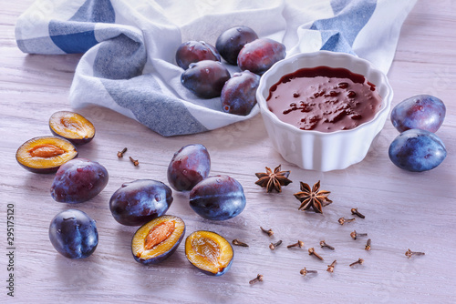 Fototapeta Naklejka Na Ścianę i Meble -  A white ceramic bowl with homemade plum jam, fresh ripe plums, crumpled towel, badyan seeds and dry carnation grains on a wooden table in the morning light