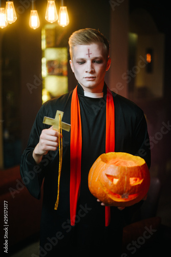 Guy in a Halloween priest costume with a serious face holds a carved gourd in his hand. The other hand raises a religious cross. A young man looks into the camera.