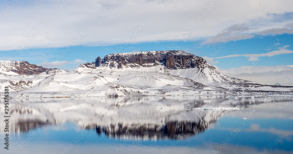 Brown hill covered with snow with reflection in still water and blue sky in Antarctica