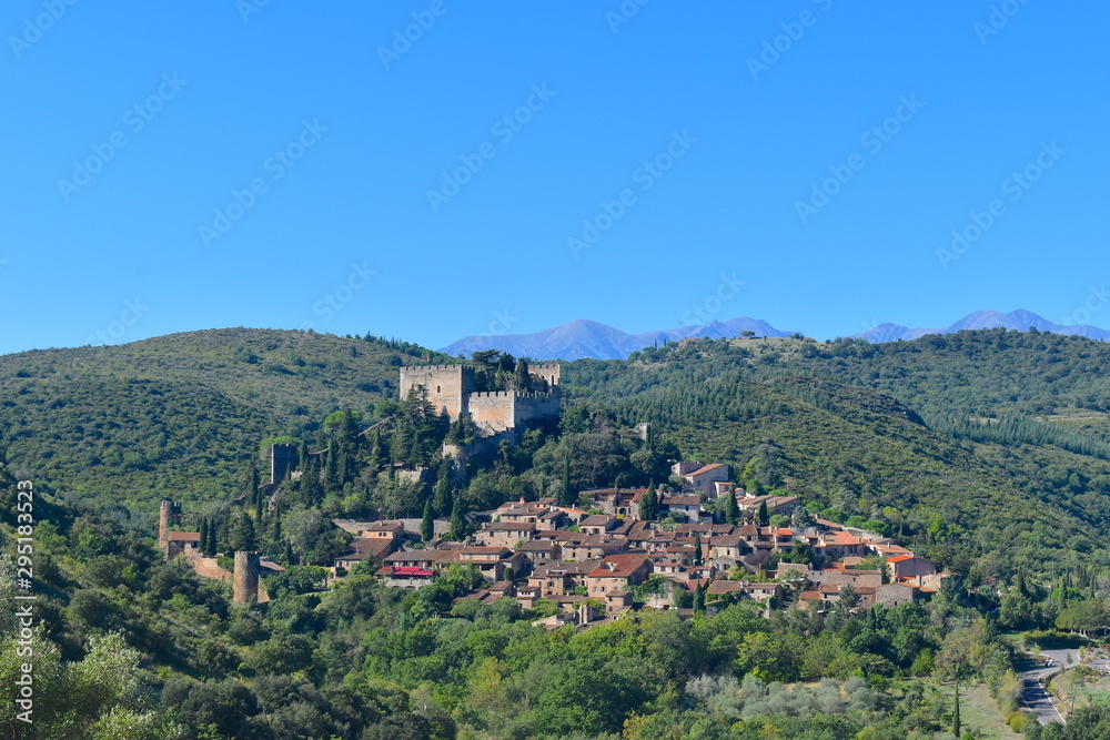 French quaint town at the foot of a castle on a hill surrounded by vineyards and greenery. Gorgeous Pyrenees mountains at the background. Castelnou, south of France