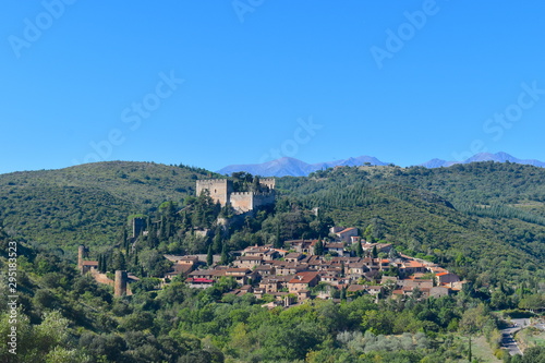 French quaint town at the foot of a castle on a hill surrounded by vineyards and greenery. Gorgeous Pyrenees mountains at the background. Castelnou, south of France