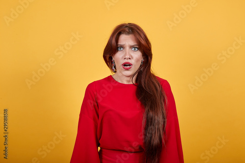 Attractive, young girl looking to the front of camera with confused face expression. Lady in red garment standing on yellow background. Brunette has brilliant, gray eyes.