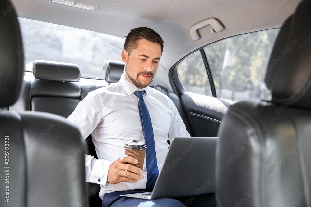 Successful businessman with laptop drinking coffee in modern car