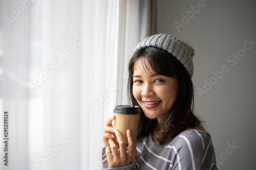 Asian woman drinking coffee in living room near the window with take away cup of coffee latte in winter time