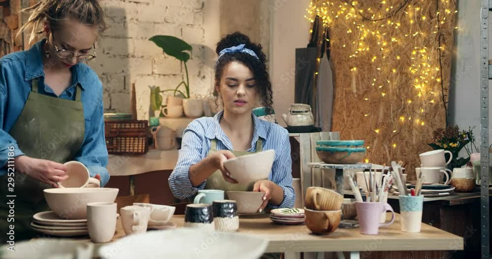 Young beautiful Caucasian girl, potter, putting away all ceramic pots and plates while making order on the desk and her friend and colleague making order at the background in the pottery studio. Young beautiful Caucasian girl, potter, putting away all ceramic pots and plates while making order on the desk and her friend and colleague making order at the background in the pottery studio.