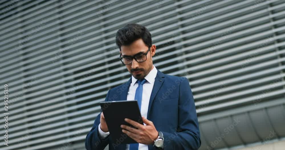 Portrait shot of the Arabian young handsome businessman in suit, tie and glasses tapping on the tablet device while standing outside and smiling to the camera. Portrait shot of the Arabian young handsome businessman in suit, tie and glasses tapping on the tablet device while standing outside and smiling to the camera.