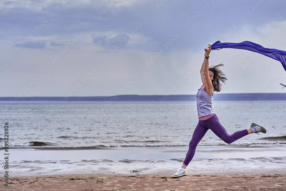 Obraz premium Young slim brunette woman jumping while jogging while holding a blue scarf in her hands. A woman is engaged in gymnastics in the spring morning on the sandy bank of a large river. Cloudy morning.